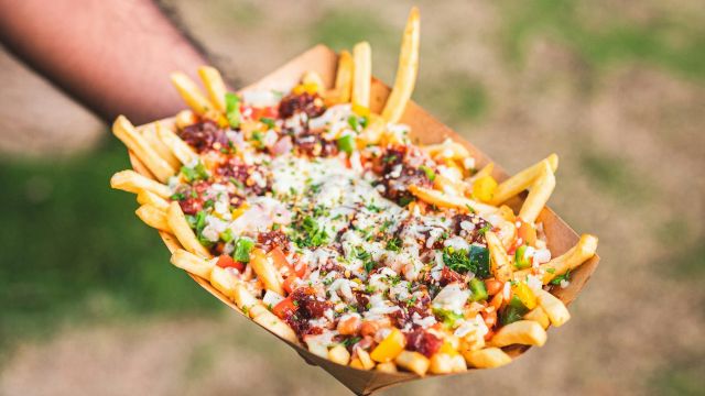 A hand holds a tray of loaded fries topped with cheese, bacon bits, and green onions over a grassy background.