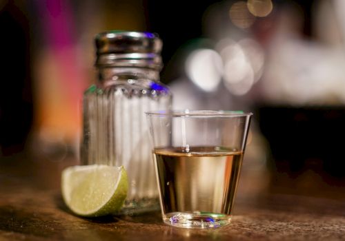 A shot glass with a golden liquid, a salt shaker, and a lime wedge on a wooden surface.