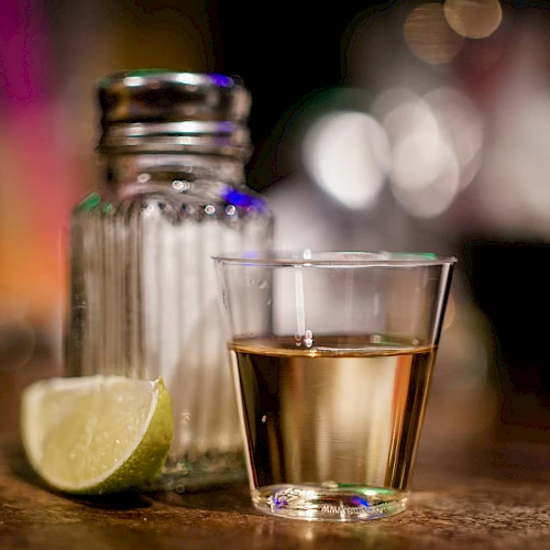 A shot glass with a golden liquid, a salt shaker, and a lime wedge on a wooden surface.