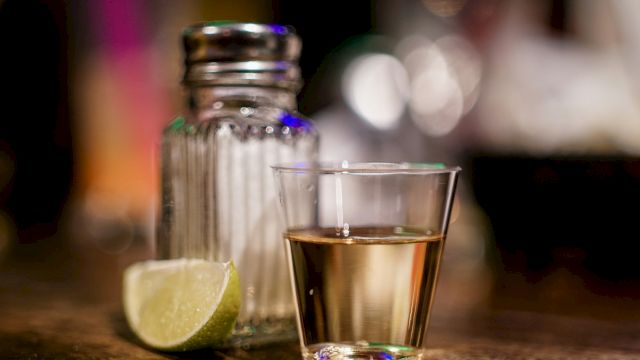 A shot glass with a golden liquid, a salt shaker, and a lime wedge on a wooden surface.