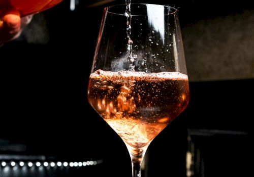A glass is being filled with a pinkish beverage, possibly rosé wine, in a dimly lit setting.