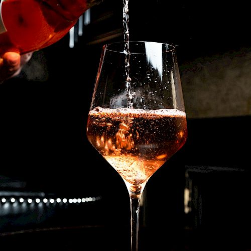 A glass is being filled with a pinkish beverage, possibly rosé wine, in a dimly lit setting.
