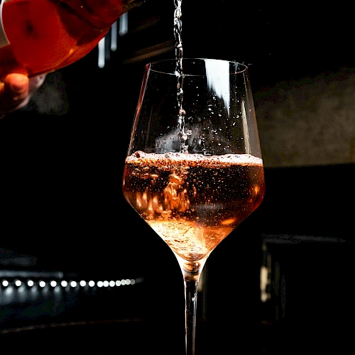 A glass is being filled with a pinkish beverage, possibly ros&eacute; wine, in a dimly lit setting.