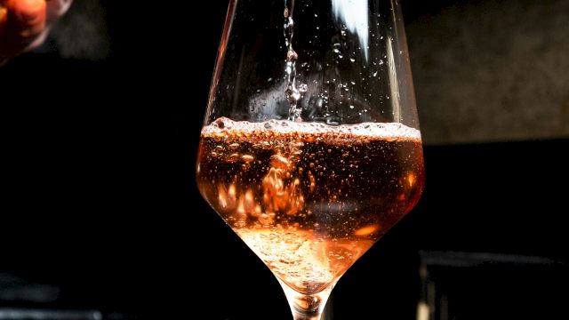 A glass is being filled with a pinkish beverage, possibly rosé wine, in a dimly lit setting.