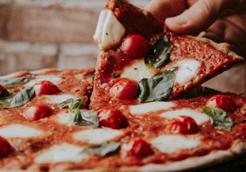 A hand lifts a slice from a Margherita pizza topped with tomatoes, mozzarella, and basil against a rustic brick wall background.