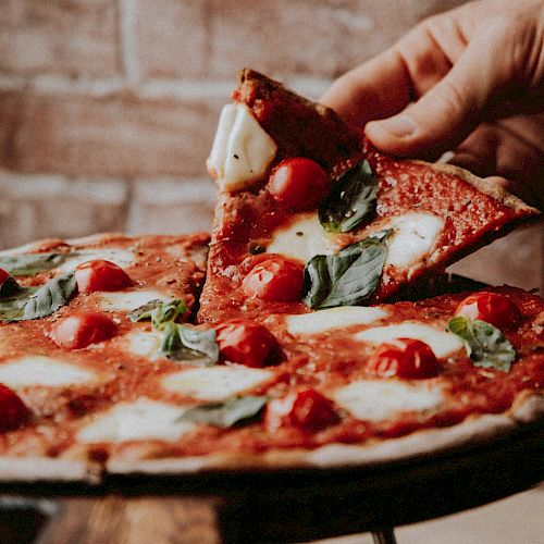 A hand lifts a slice from a Margherita pizza topped with tomatoes, mozzarella, and basil against a rustic brick wall background.