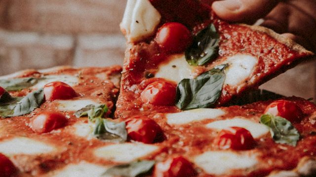 A hand lifts a slice from a Margherita pizza topped with tomatoes, mozzarella, and basil against a rustic brick wall background.