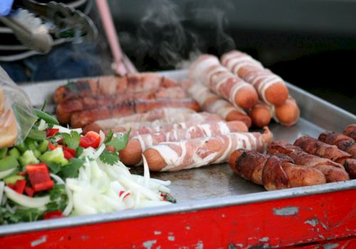 The image shows a grill with bacon-wrapped hot dogs and assorted vegetables, including onions and bell peppers, on a metal tray.
