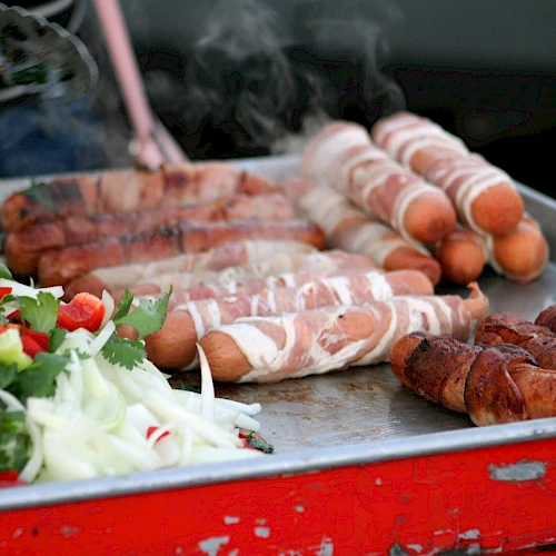 The image shows a grill with bacon-wrapped hot dogs and assorted vegetables, including onions and bell peppers, on a metal tray.