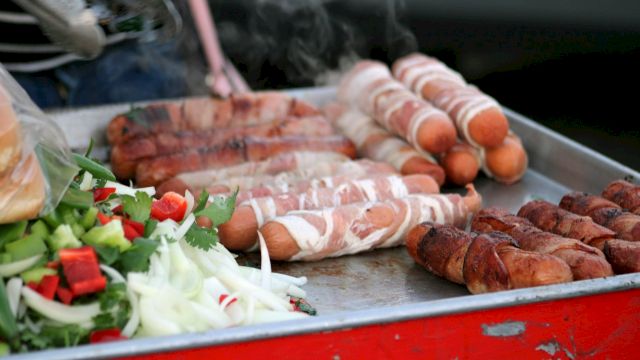The image shows a grill with bacon-wrapped hot dogs and assorted vegetables, including onions and bell peppers, on a metal tray.