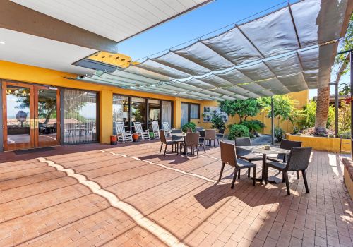 Outdoor patio with brick flooring, tables, and chairs, shaded by a large canopy, adjacent to a building with glass doors and yellow walls.