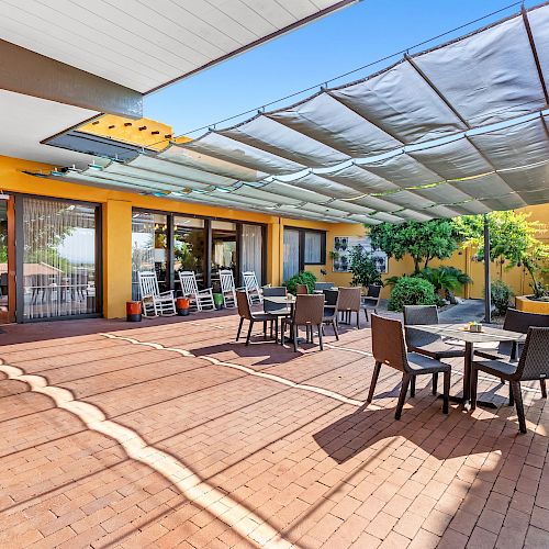 Outdoor patio with brick flooring, tables, and chairs, shaded by a large canopy, adjacent to a building with glass doors and yellow walls.