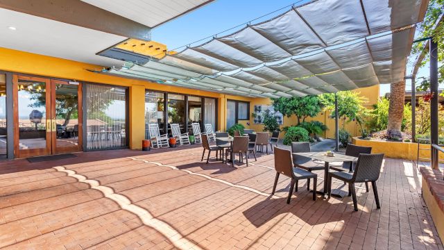 Outdoor patio with brick flooring, tables, and chairs, shaded by a large canopy, adjacent to a building with glass doors and yellow walls.