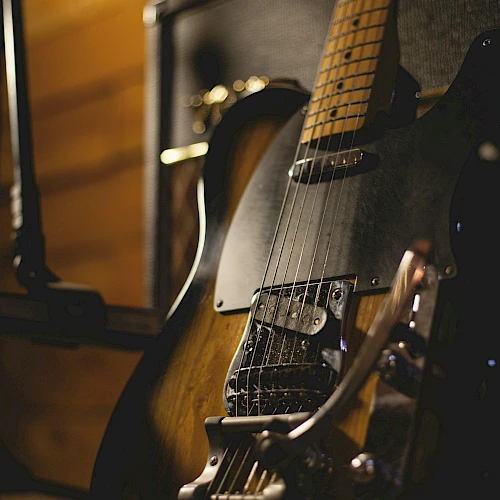 The image shows an electric guitar resting against an amplifier in a dimly lit setting, showcasing its wooden body and strings.