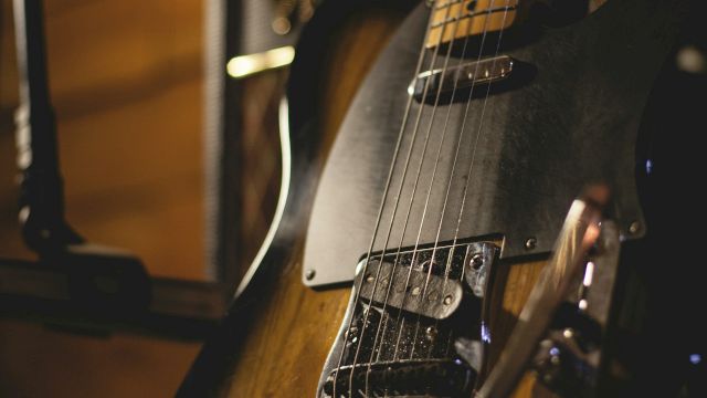 The image shows an electric guitar resting against an amplifier in a dimly lit setting, showcasing its wooden body and strings.