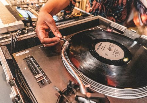 A person is adjusting a needle on a turntable playing a vinyl record, surrounded by audio equipment in a DJ setup.