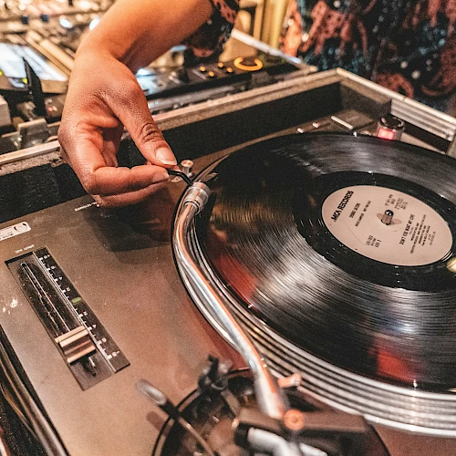 A person is adjusting a needle on a turntable playing a vinyl record, surrounded by audio equipment in a DJ setup.
