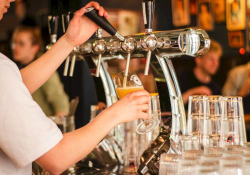 A person is pouring draft beer into a glass at a bar, with rows of empty glasses visible in the foreground.