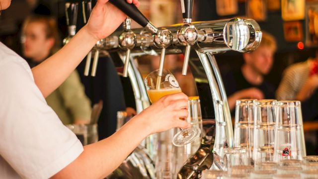 A person is pouring draft beer into a glass at a bar, with rows of empty glasses visible in the foreground.