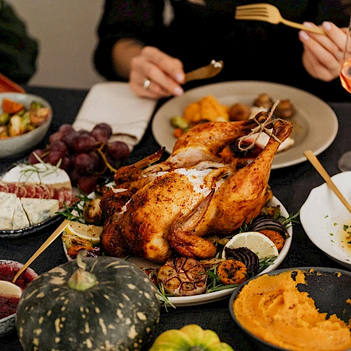 A table with a roasted turkey, various sides like mashed sweet potatoes and brussels sprouts, wine glasses, and apples is shown.
