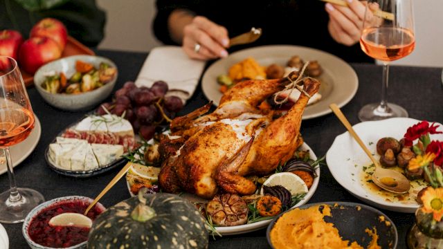 A table with a roasted turkey, various sides like mashed sweet potatoes and brussels sprouts, wine glasses, and apples is shown.