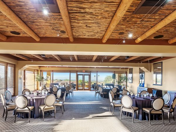 A bright banquet hall with round tables and chairs arranged for a reception, large windows and doorways at the far end, wooden ceiling beams, and a cozy, airy setup.
