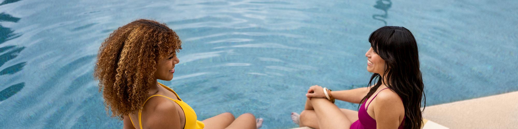Two women in swimsuits sitting by a pool, chatting near the water.