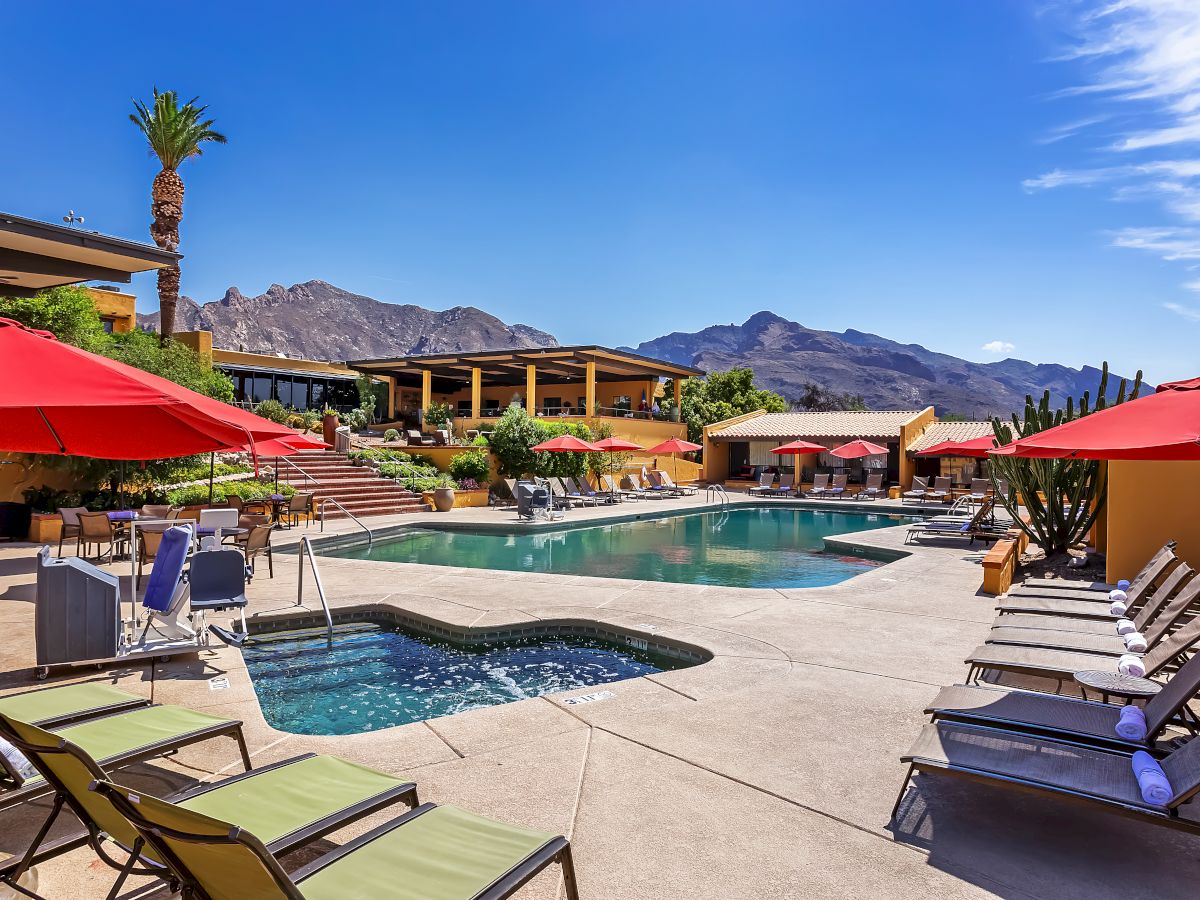 A sunny resort pool area with lounge chairs, red umbrellas, a hot tub, and mountains in the background, under a clear blue sky.
