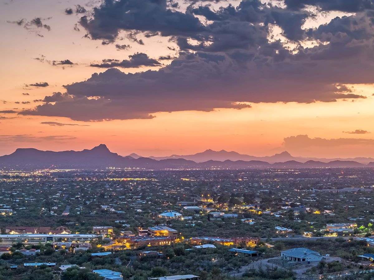 A desert city skyline at dusk with a warm orange glow, distant purple mountains, scattered lights below, and dramatic clouds overhead.