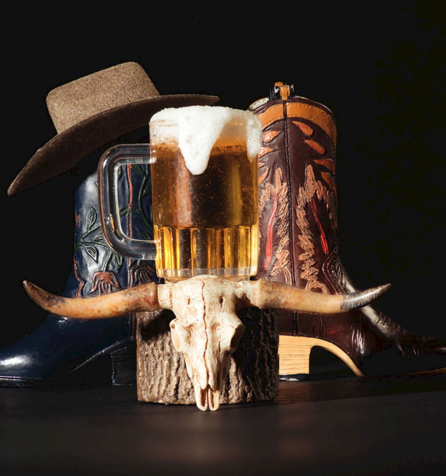 A beer mug perched on a rustic skull with horns, cowboy hat, boot, and rustic wood accents, all set against a dark backdrop.
