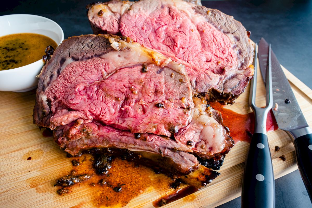 Juicy medium-rare roast beef slices on a wooden board, with carving knives and a bowl of sauce.