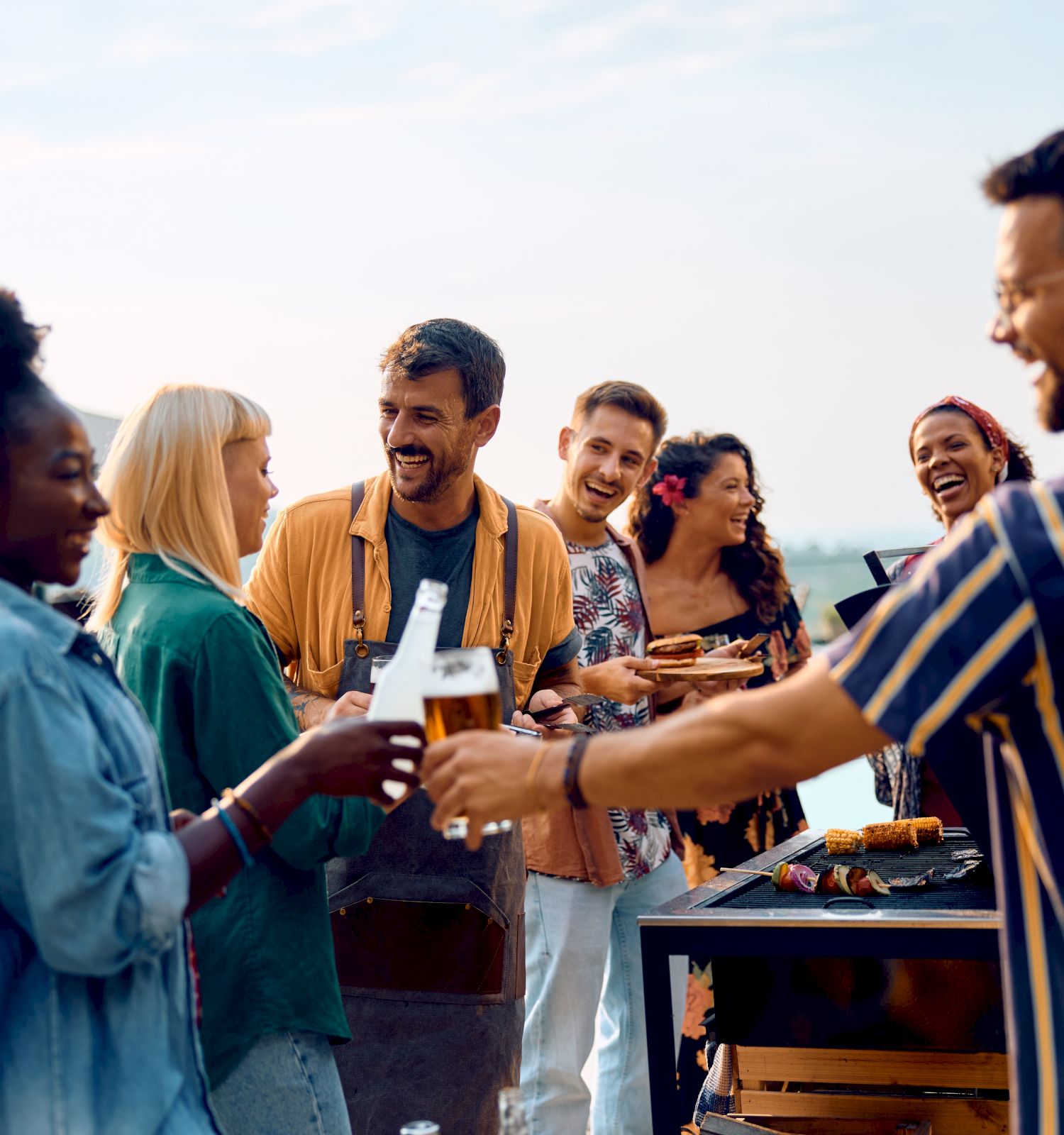 A group of friends at an outdoor barbecue, laughing as they serve food from a grill under a sunny sky.
