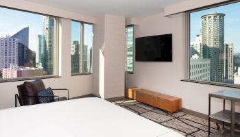 A modern hotel room with large windows offering a city view, a bed, a chair, a TV on the wall, and a wooden console below it.