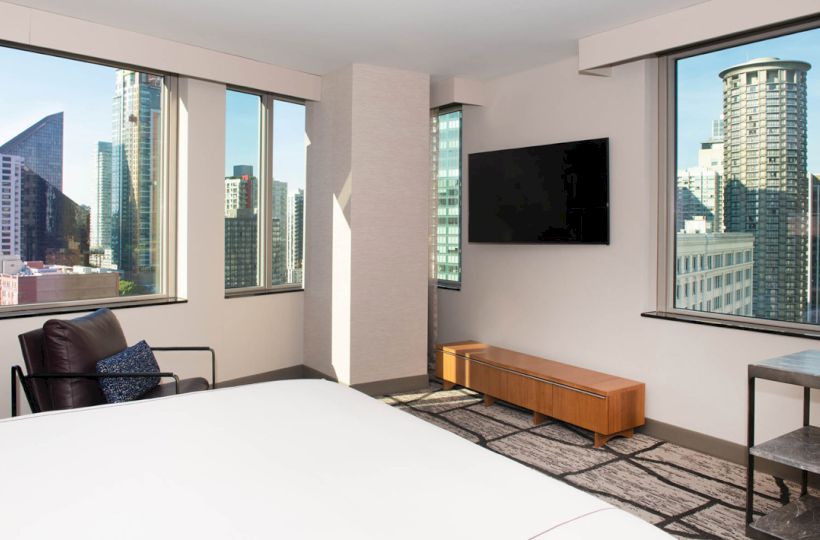 A modern hotel room with large windows offering a city view, a bed, a chair, a TV on the wall, and a wooden console below it.