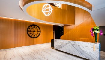 A modern lobby with a spiral stairway, a large wall clock, marble reception desk, and a flower vase.