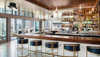 A modern bar with a wooden countertop, five barstools, a well-stocked liquor shelf, and large windows offering city views, under a sleek ceiling.