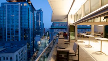 A modern rooftop balcony at dusk with seating areas, cityscape view, and outdoor tables, overlooking downtown high-rise buildings and a well-lit street.