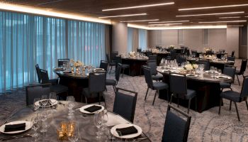 A banquet hall with round tables set for a formal event, featuring black chairs, white floral centerpieces, and dim, modern lighting.