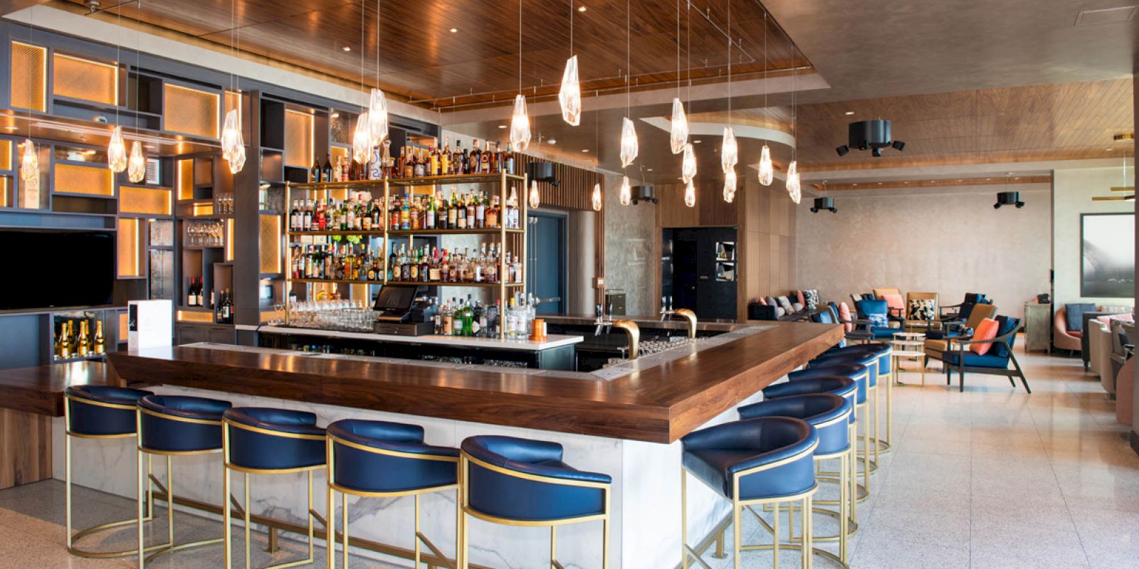 The image shows a modern bar with high chairs, hanging lights, and a wide selection of bottles on shelves behind the counter, with seating areas.