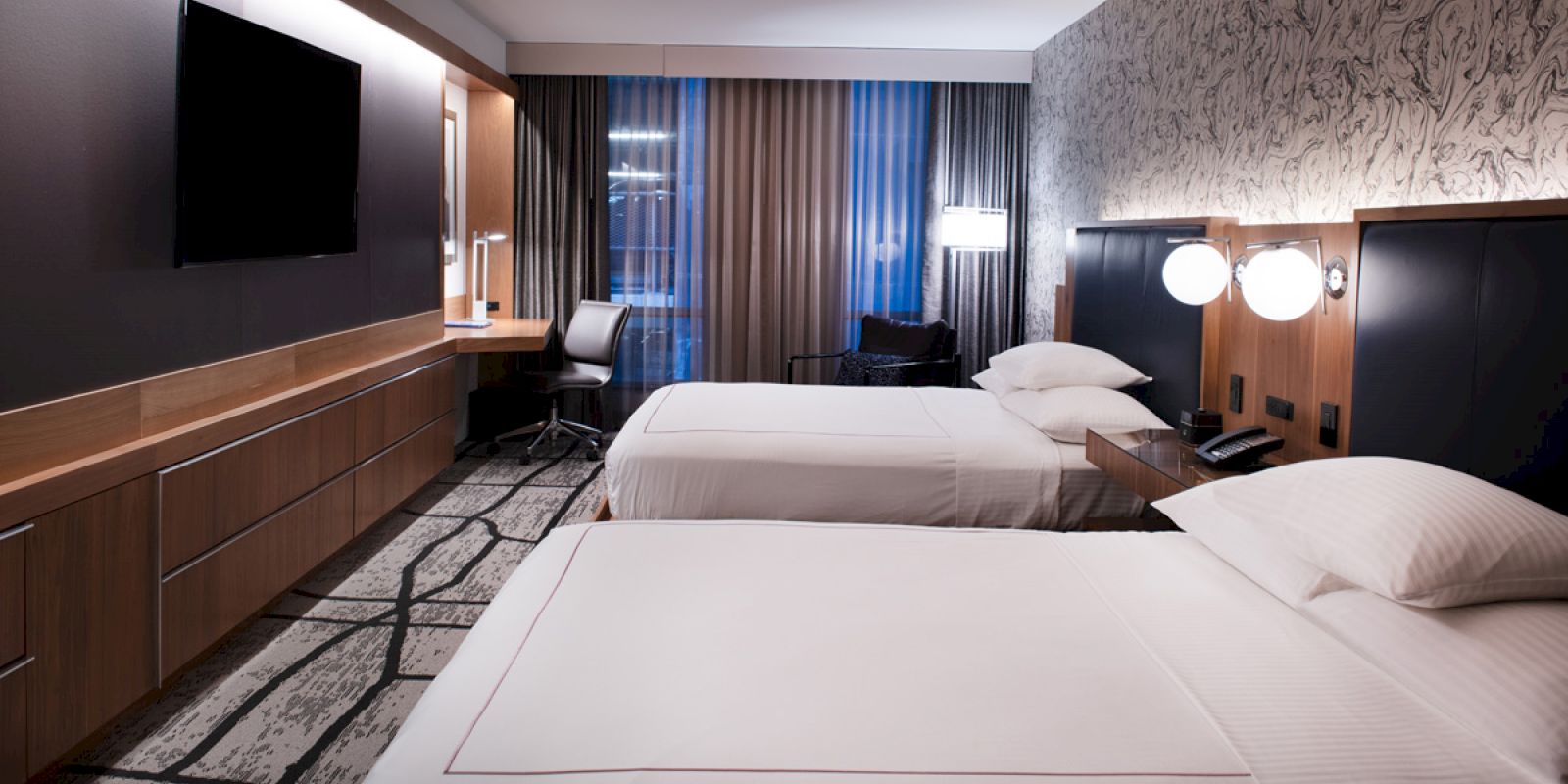 This image shows a modern hotel room with two neatly made beds, a large TV, a desk with a chair, and stylish decor.