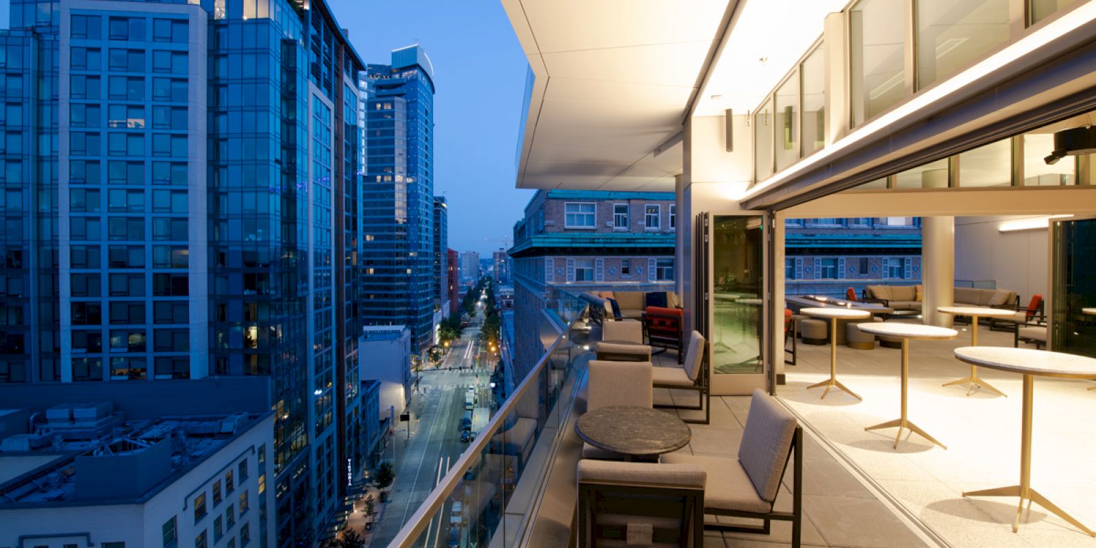 This image shows a modern balcony overlooking a cityscape with high-rise buildings at dusk. The balcony has tables and chairs.
