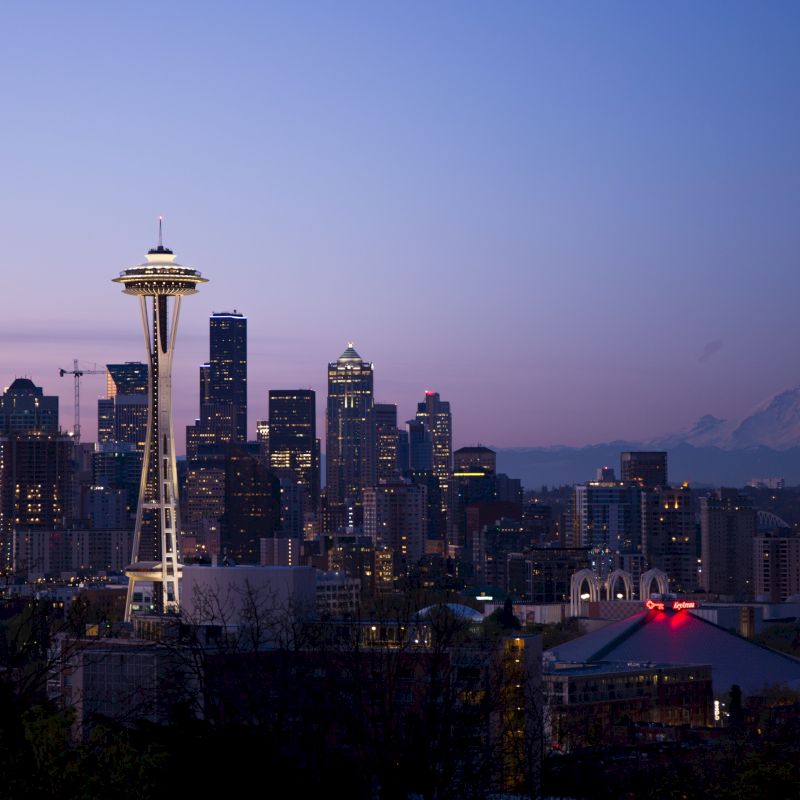 The image shows a cityscape at dusk featuring a prominent tower, high-rise buildings, and a distant mountain under a twilight sky.
