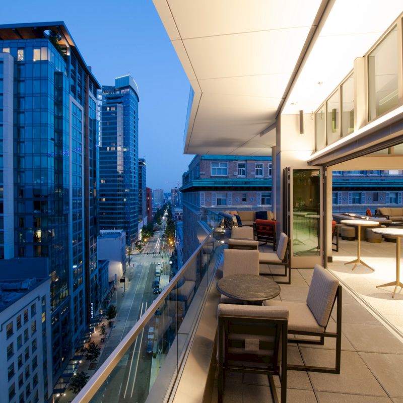 A modern balcony view overlooking a cityscape at dusk, featuring outdoor seating and tables, with adjacent buildings illuminated by city lights.
