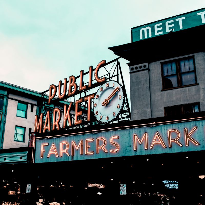 This image shows a public market with a large clock and neon sign reading "Public Market" and "Farmers Market" against the backdrop of building façades.