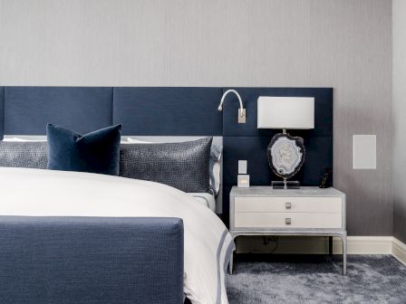 A modern bedroom featuring a bed with blue and white bedding, a blue cushioned headboard, a nightstand with a lamp and decorative items.
