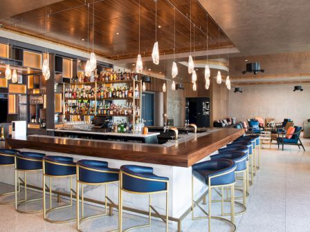 This image shows a modern bar with a wooden counter, blue and gold bar stools, hanging lights, and shelves stocked with bottles.