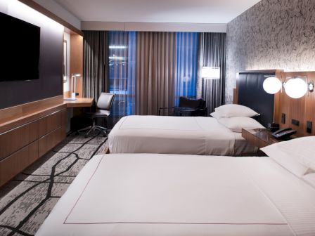 A modern hotel room with two neatly made beds, a television on the wall, a desk with a chair, and stylish decor, including a patterned wall and curtains.