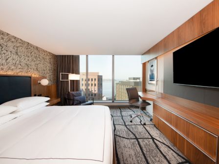 A modern hotel room with a large bed, wall-mounted TV, desk, chair, and a large window offering a view of buildings and water in the distance.