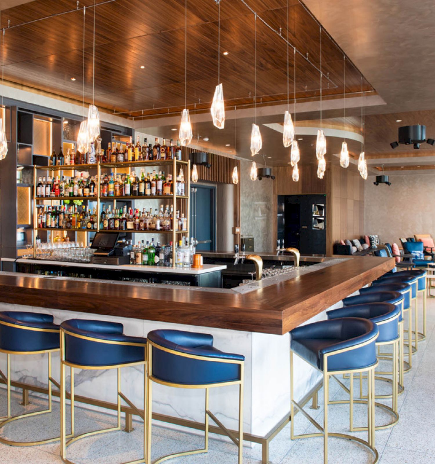 The image shows a modern bar with high stools, a wooden counter, hanging lights, and shelves stocked with various bottles behind the bar.