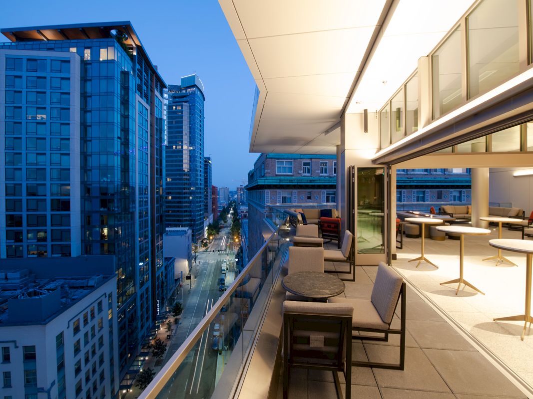 The image shows a modern cityscape at dusk with tall buildings and a spacious balcony area furnished with tables and chairs.